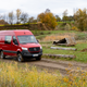 a red 2023 mercedes benz sprinter awd van driving on a dirt trail through a field, with autumn trees and rolling hills in the background